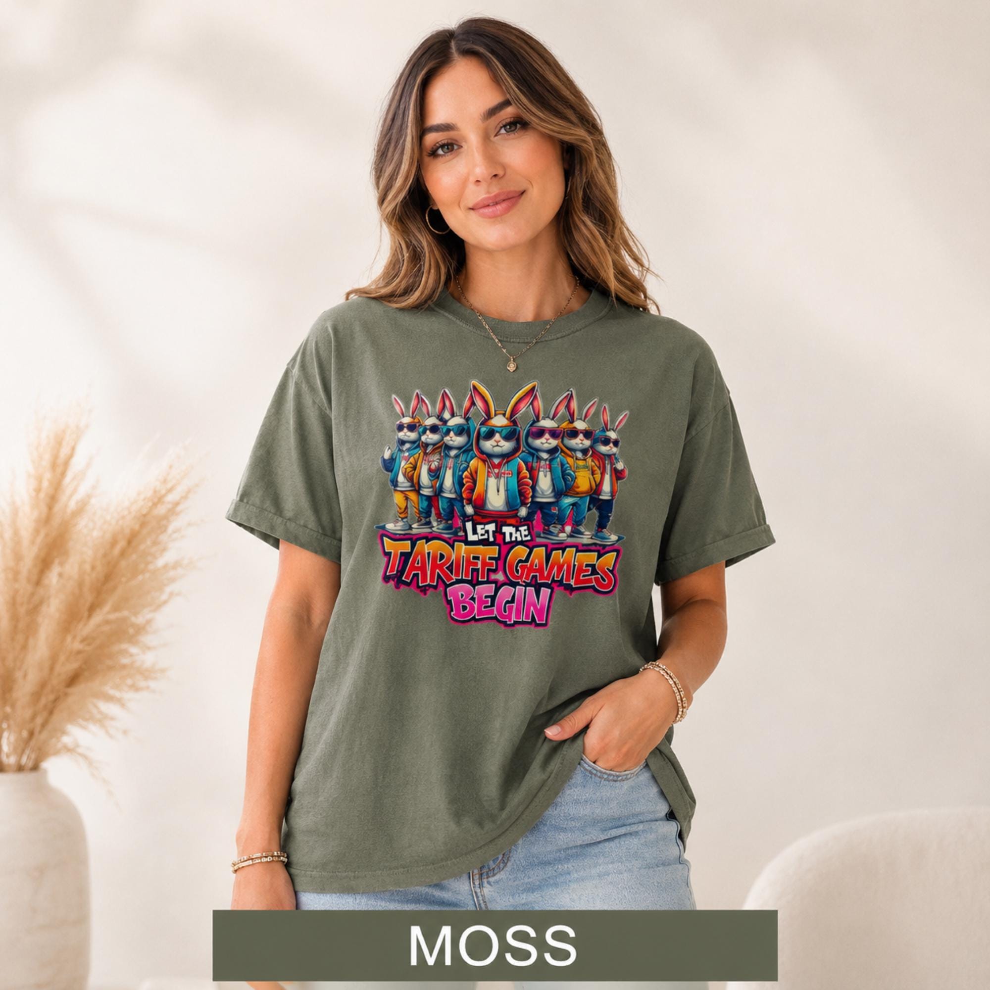 Female model wearing a moss green T-shirt featuring stylish cartoon rabbits and bold “Let the Tariff Games Begin” text.