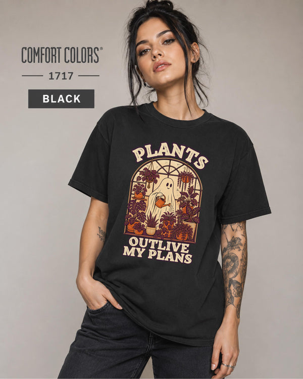 Model wearing Comfort colors tshirt in the color black featuring a Retro cream and orange ghost watering purple houseplants inside a greenhouse with the words Plants Outlive My Plans.