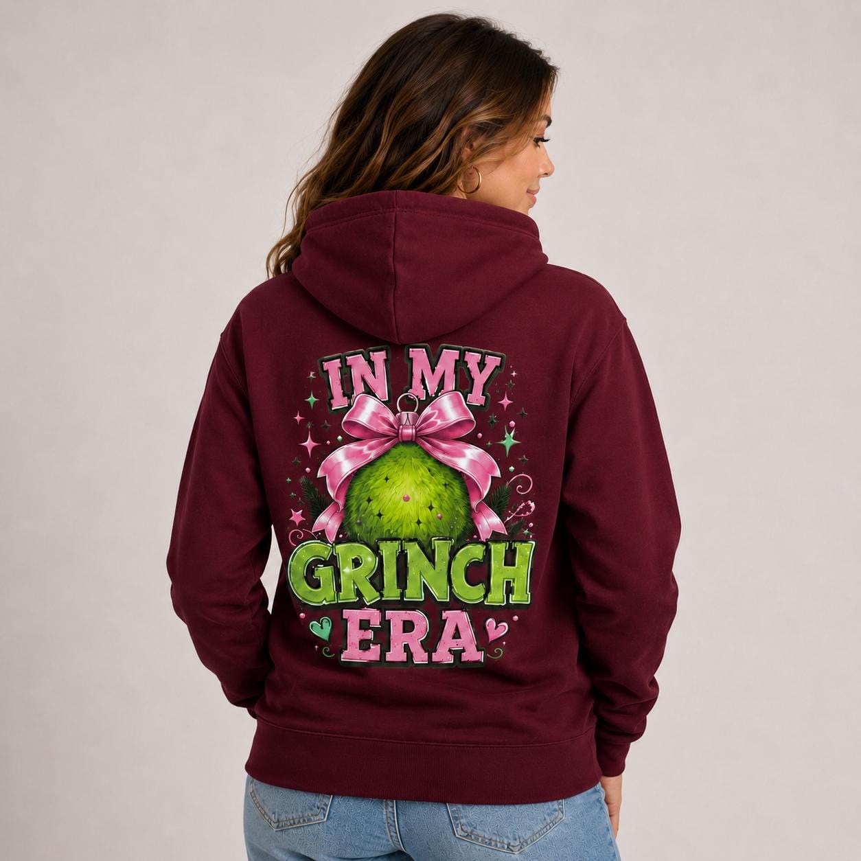 Female model wearing a maroon holiday hoodie with a pink and green festive character design printed on the front.