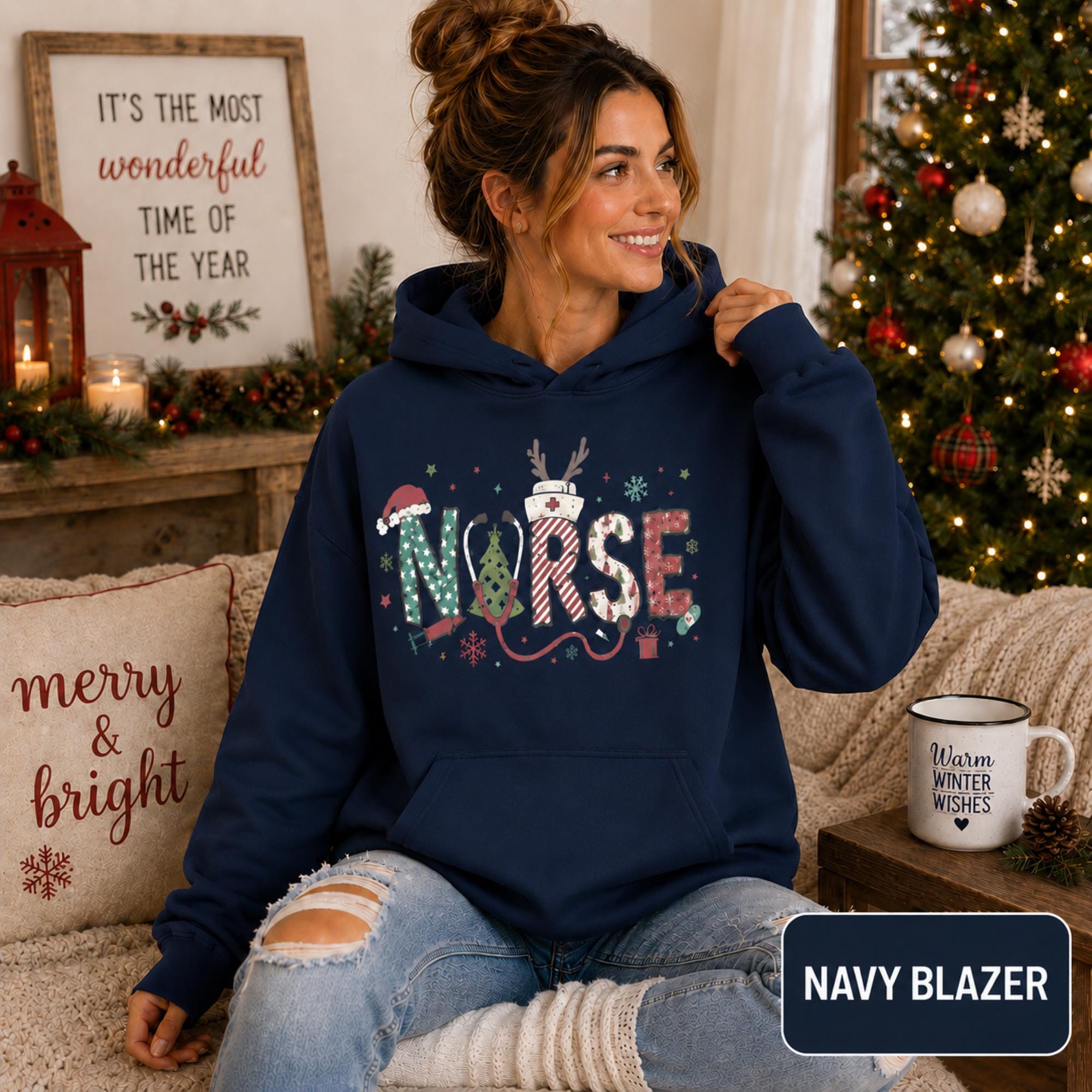 Female model wearing a navy blazer Christmas nurse hoodie with colorful festive NURSE lettering in a cozy holiday living room.
