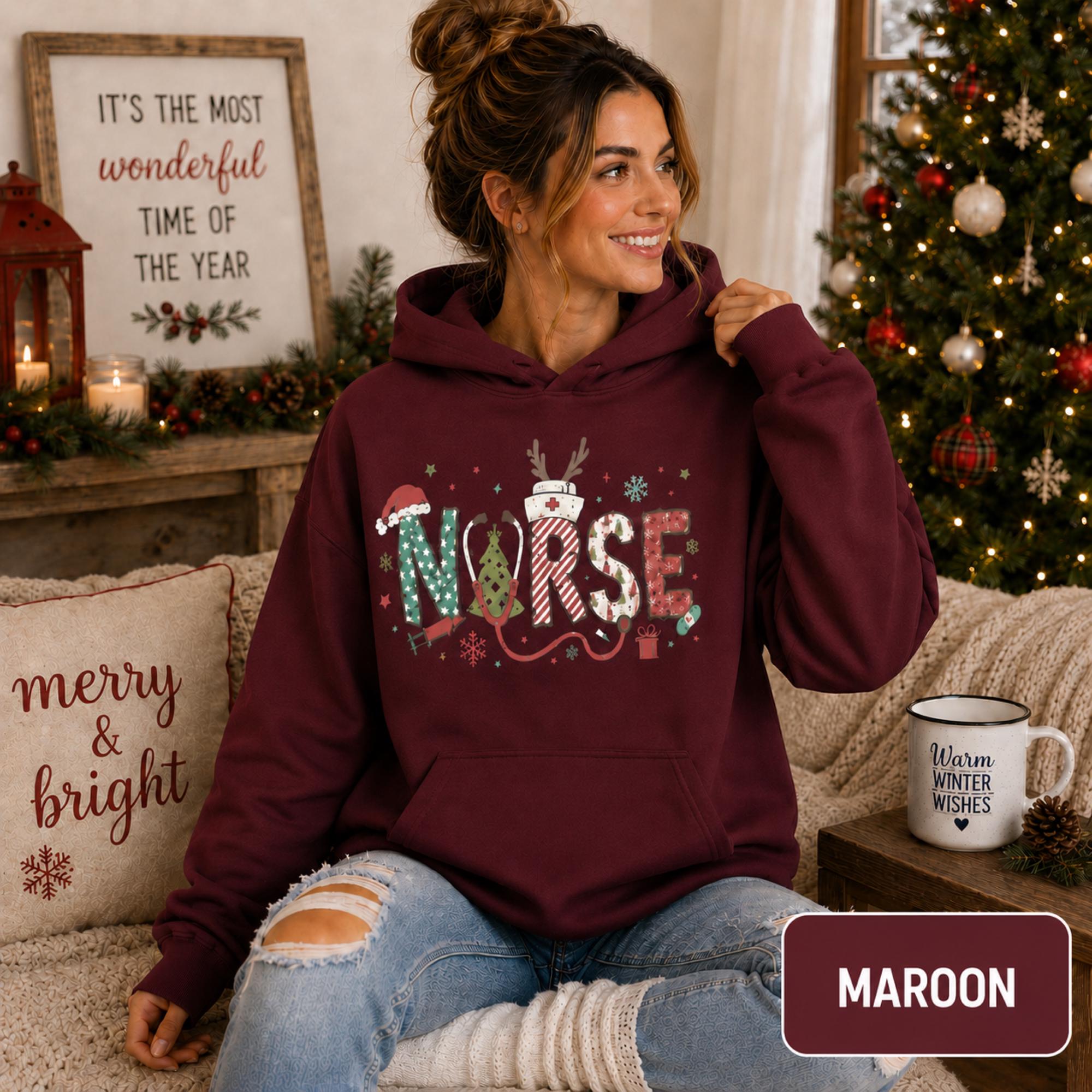 Female model wearing a maroon Christmas nurse hoodie with colorful festive NURSE lettering in a cozy holiday living room.