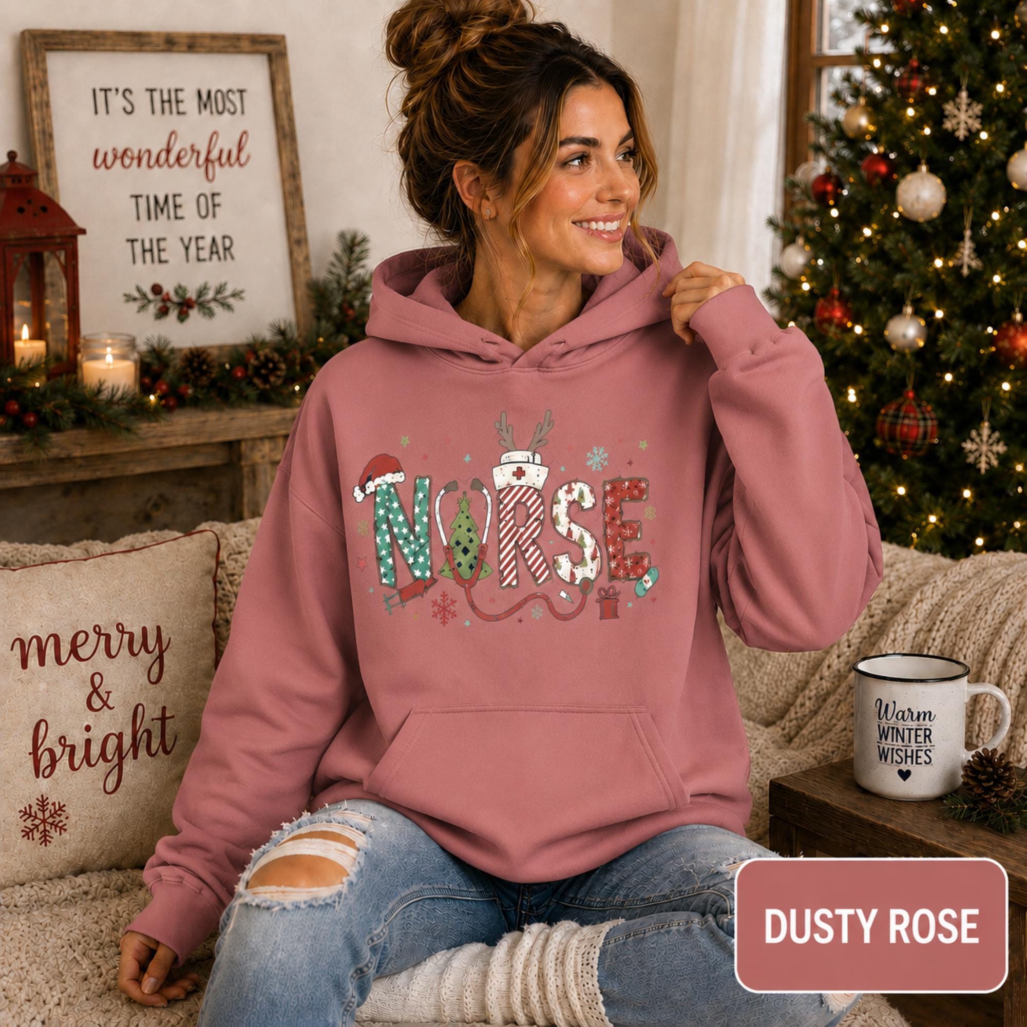 Female model wearing a dusty rose Christmas nurse hoodie with colorful festive NURSE lettering in a cozy holiday living room.
