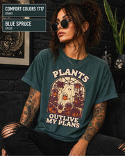 Model wearing Comfort colors tshirt in the color blue spruce featuring a Retro cream and orange ghost watering purple houseplants inside a greenhouse with the words Plants Outlive My Plans.