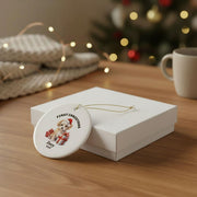 Personalized First Christmas ceramic ornament displayed beside a white gift box on a wooden table, featuring a festive Santa design and custom name in an elegant holiday gift setting.