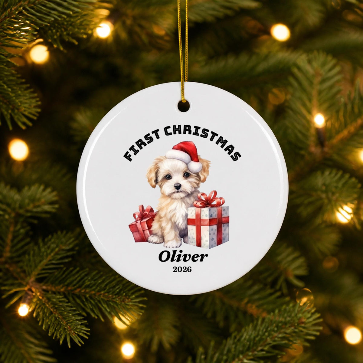 Close-up of a personalized First Christmas ornament hanging from a Christmas tree with golden fairy lights, showing festive Santa artwork, custom name Oliver, and a white circular ceramic finish.
