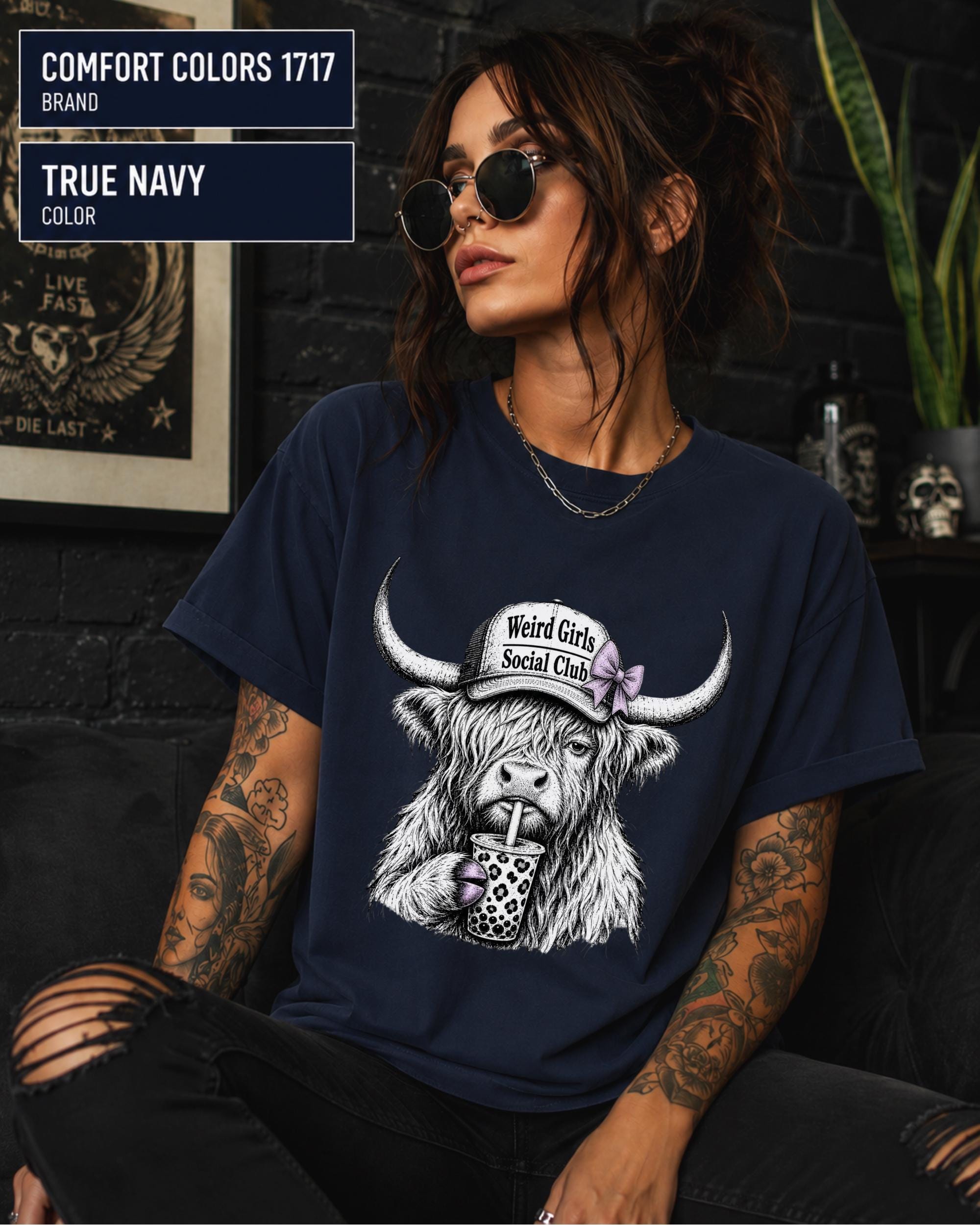 True navy graphic tee with a shaggy Highland cow, lavender bow, trucker cap, and spotted beverage cup.