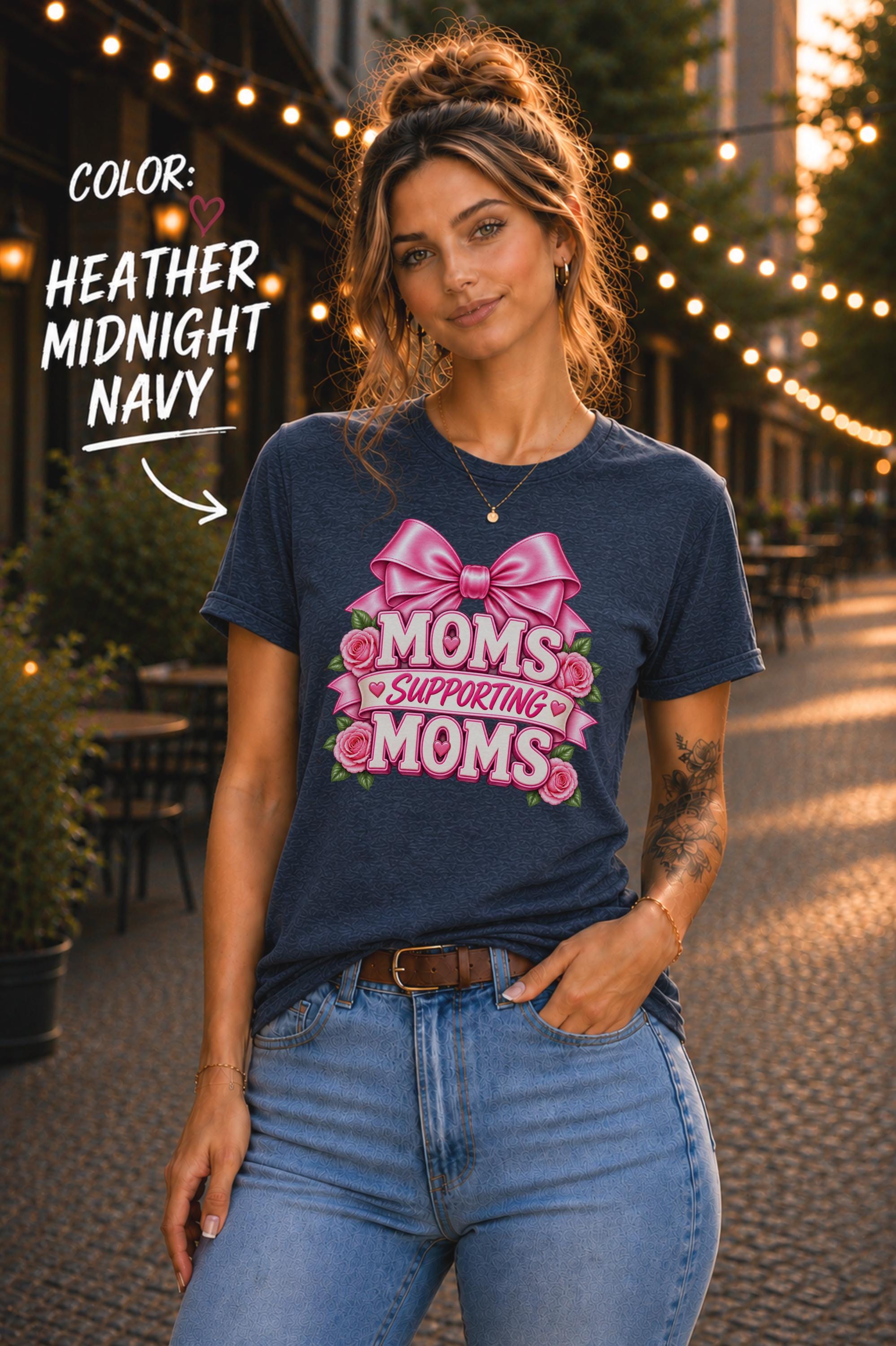 Woman wearing a Heather Midnight Navy graphic T-shirt featuring the phrase “Moms Supporting Moms” in pink lettering with a large pink bow, roses, and heart accents, styled with light-wash jeans in an outdoor café setting with warm string lights.