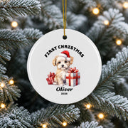 Personalized white ceramic First Christmas ornament hanging from a snowy evergreen tree, featuring a festive Santa illustration, custom name Oliver, and warm holiday lights.