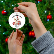 Hands hanging a personalized First Christmas ornament on a decorated green Christmas tree, featuring festive Santa artwork, the name Oliver, and a white round ceramic design.