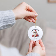 Close-up of hands holding a personalized First Christmas ceramic ornament by its hanging cord, showing the round shape, festive Santa illustration, and custom name Oliver.