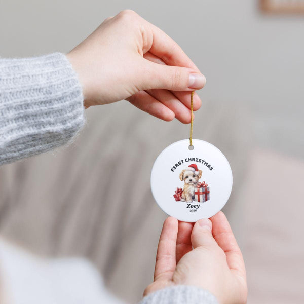 Close-up of hands holding a personalized First Christmas ceramic ornament by its hanging cord, showing the round shape, festive Santa illustration, and custom name Oliver.