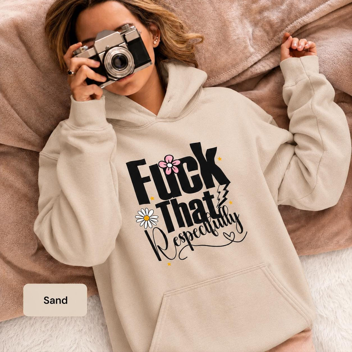 Woman wearing a sand Gildan 18500 hoodie with “Fuck That Respectfully” graphic, daisy accents, script text, and warm neutral blanket styling.