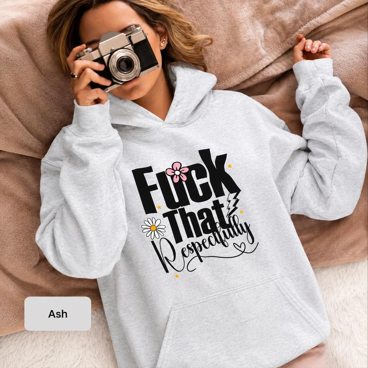 Woman wearing an ash Gildan 18500 hoodie featuring a bold “Fuck That Respectfully” floral graphic while holding a vintage camera on a cozy neutral blanket.