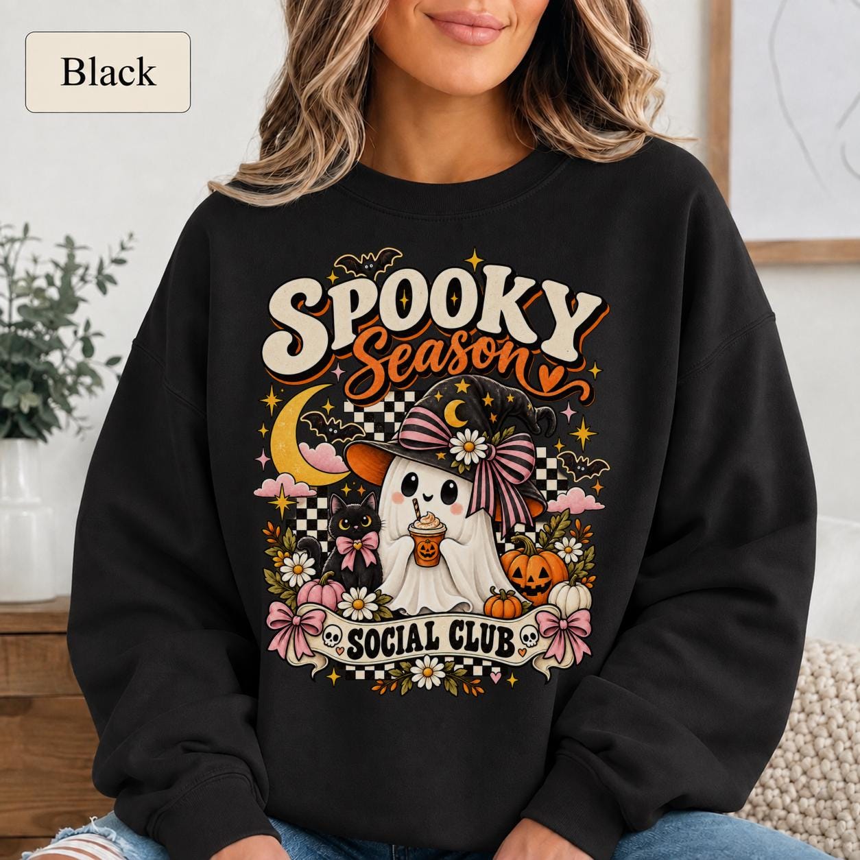 Woman wearing a black Spooky Season Social Club sweatshirt with ghost, black cat, pumpkins, flowers, and pink bows.