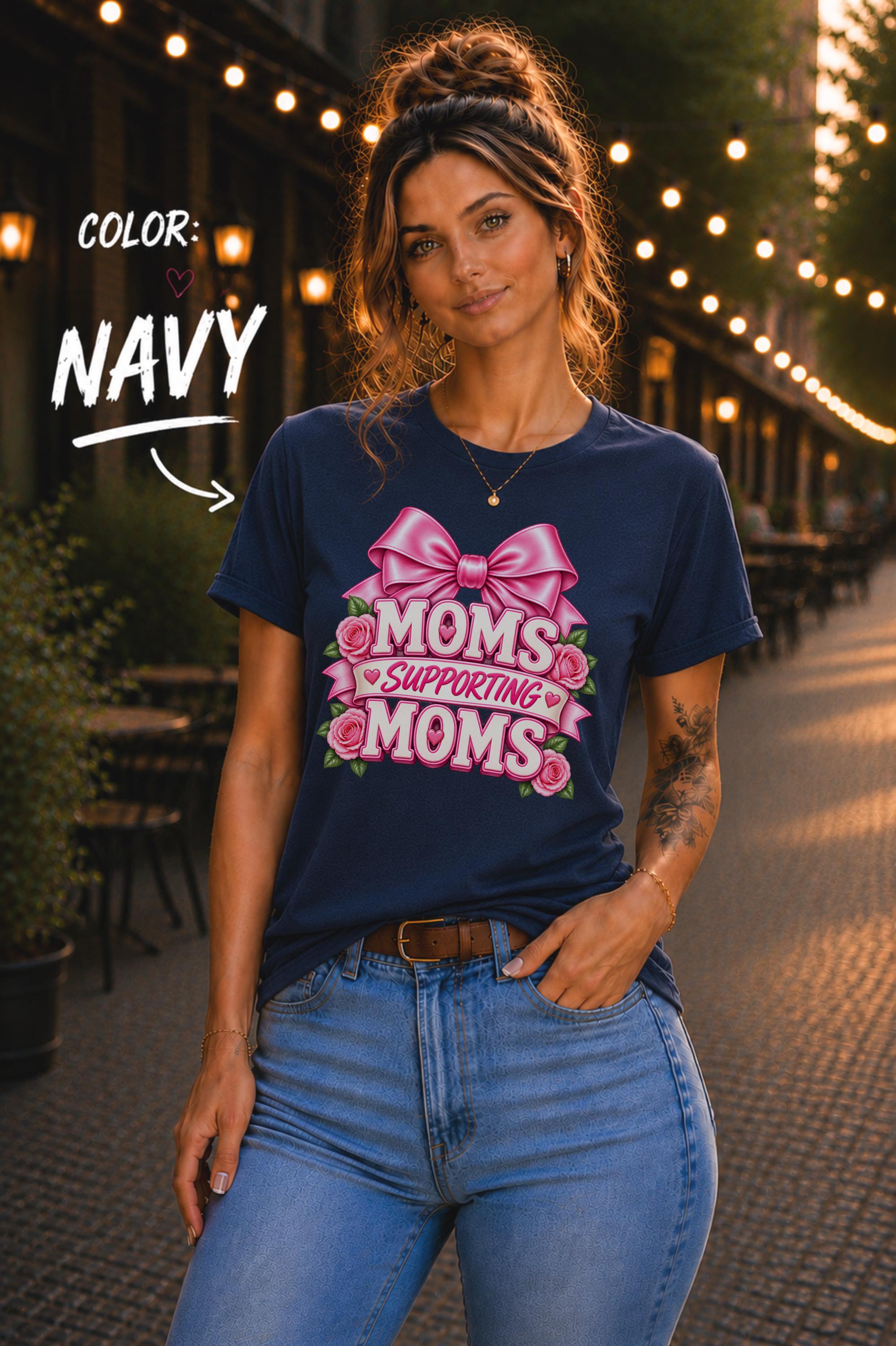 Woman wearing a Navy graphic T-shirt featuring the phrase “Moms Supporting Moms” in pink lettering with a large pink bow, roses, and heart accents, styled with light-wash jeans in an outdoor café setting with warm string lights.