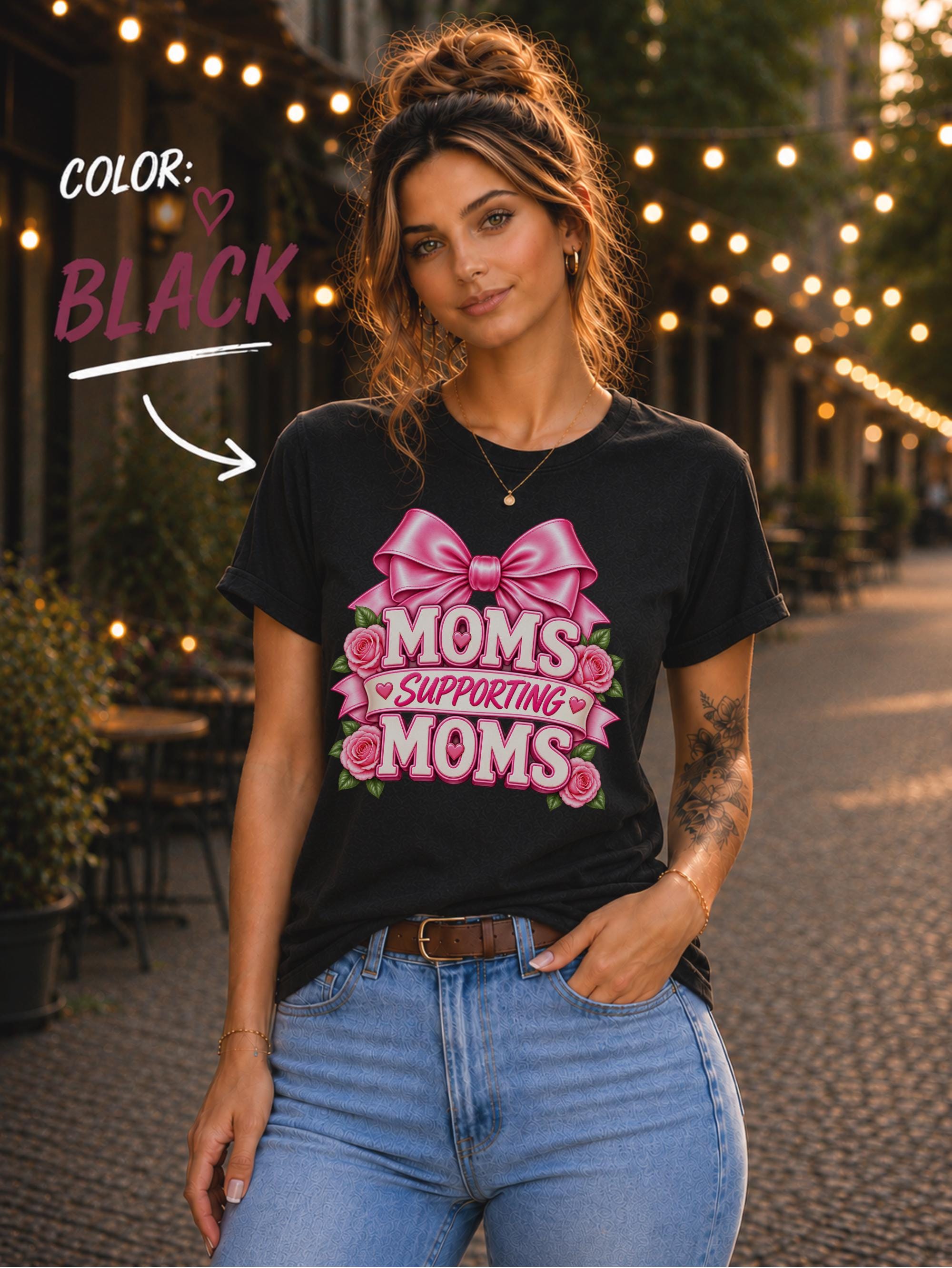 Woman wearing a Black graphic T-shirt featuring the phrase “Moms Supporting Moms” in pink lettering with a large pink bow, roses, and heart accents, styled with light-wash jeans in an outdoor café setting with warm string lights.