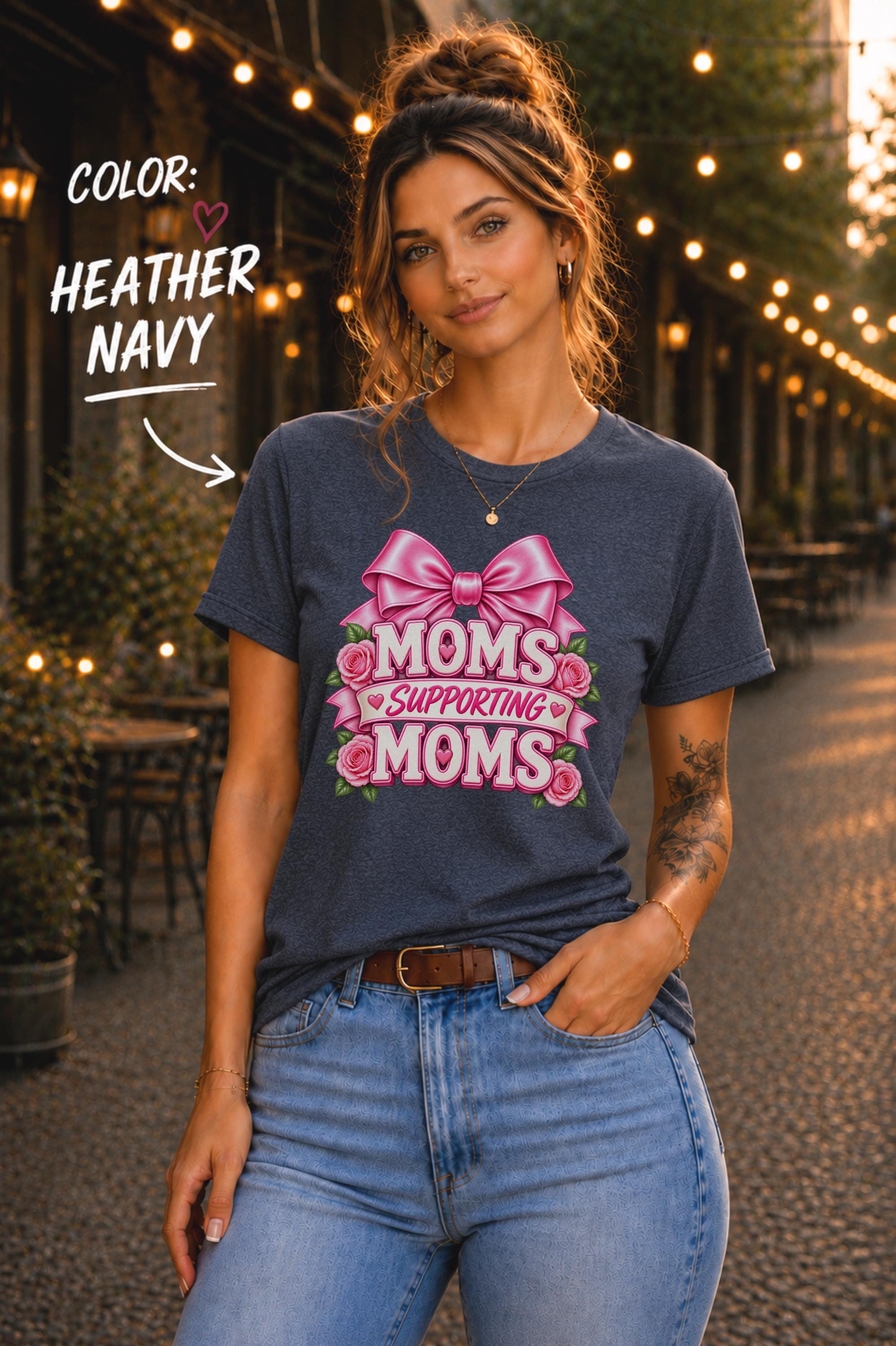 Woman wearing a Heather Navy graphic T-shirt featuring the phrase “Moms Supporting Moms” in pink lettering with a large pink bow, roses, and heart accents, styled with light-wash jeans in an outdoor café setting with warm string lights.