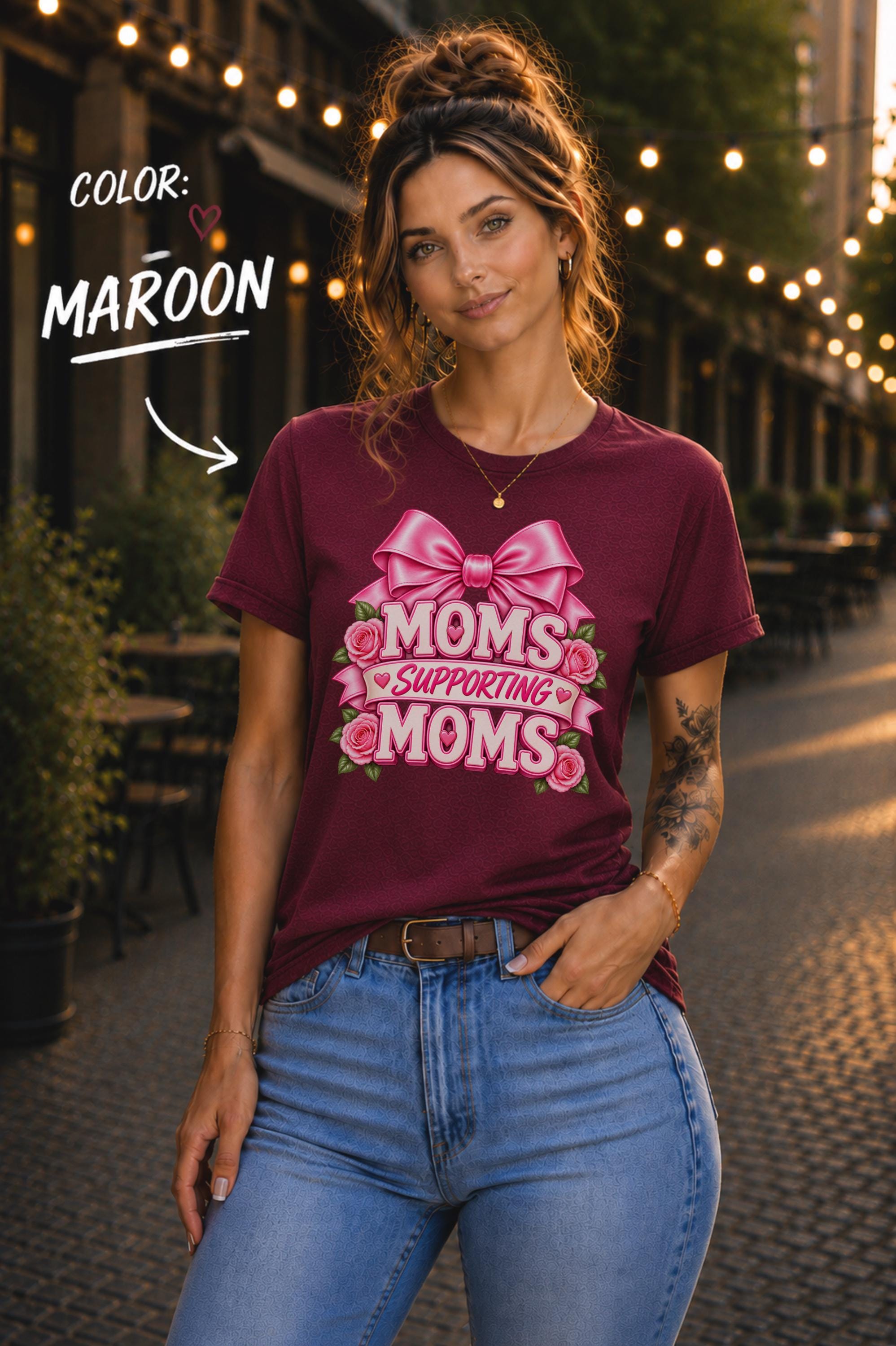 Woman wearing a Maroon graphic T-shirt featuring the phrase “Moms Supporting Moms” in pink lettering with a large pink bow, roses, and heart accents, styled with light-wash jeans in an outdoor café setting with warm string lights.
