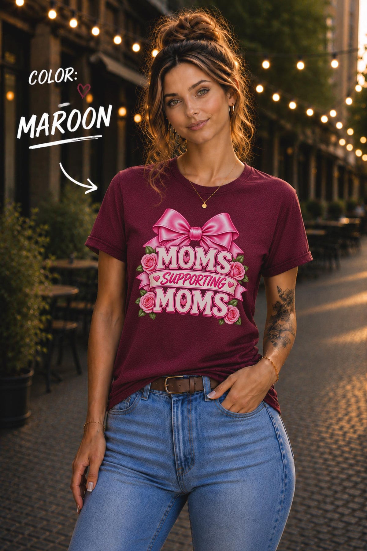 Woman wearing a Maroon graphic T-shirt featuring the phrase “Moms Supporting Moms” in pink lettering with a large pink bow, roses, and heart accents, styled with light-wash jeans in an outdoor café setting with warm string lights.