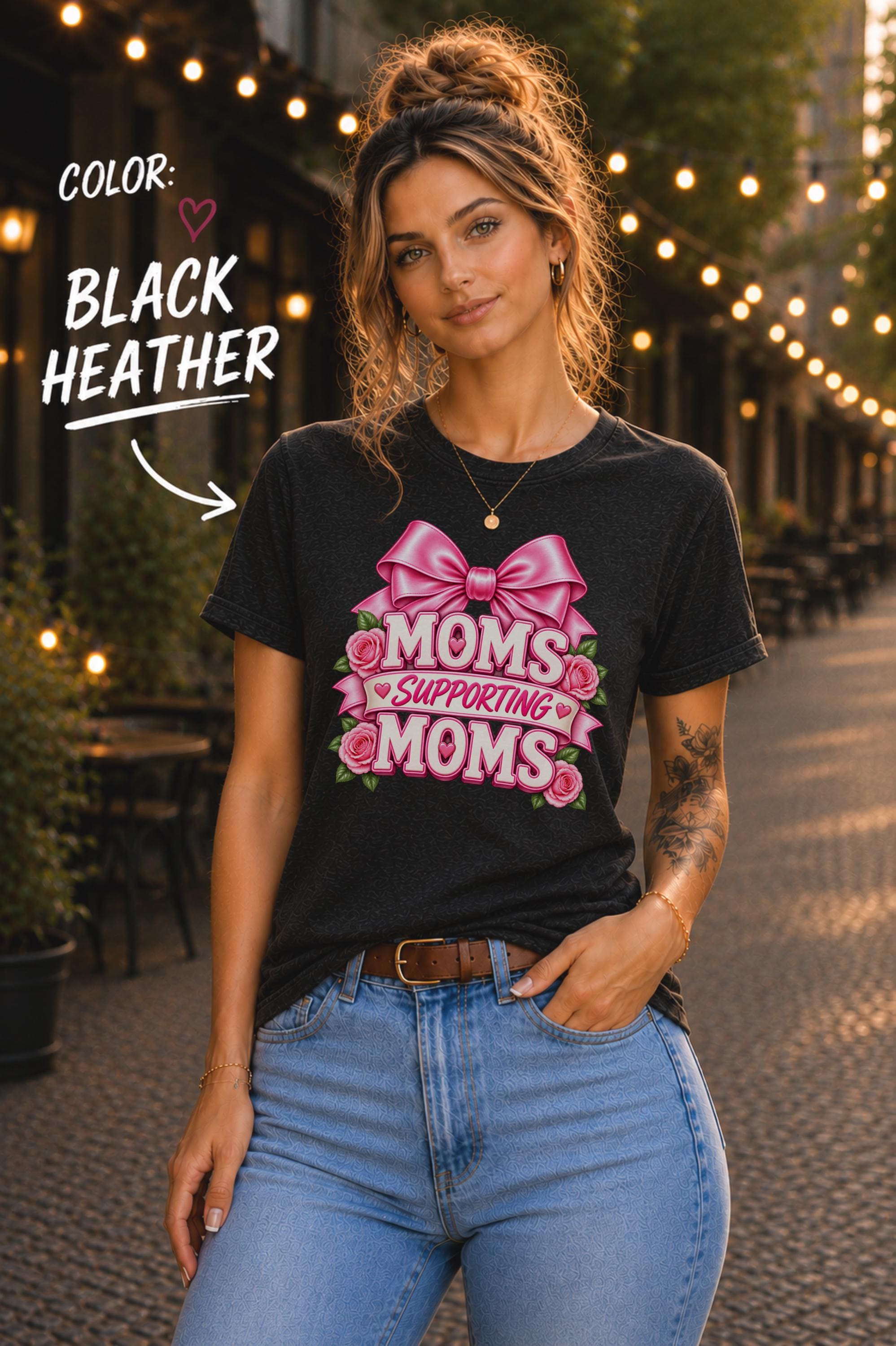 Woman wearing a Black Heather graphic T-shirt featuring the phrase “Moms Supporting Moms” in pink lettering with a large pink bow, roses, and heart accents, styled with light-wash jeans in an outdoor café setting with warm string lights.