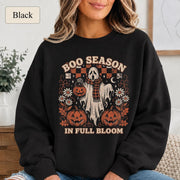 Woman wearing a black Boo Season in Full Bloom Halloween sweatshirt with ghost, pumpkins, flowers, and retro artwork.