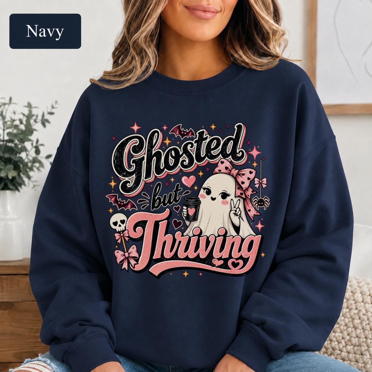 Woman wearing a navy crewneck sweatshirt featuring a cute bow-wearing ghost holding coffee, surrounded by pink hearts, bats, stars, and the words Ghosted but Thriving.