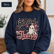 Woman wearing a navy crewneck sweatshirt featuring a cute bow-wearing ghost holding coffee, surrounded by pink hearts, bats, stars, and the words Ghosted but Thriving.