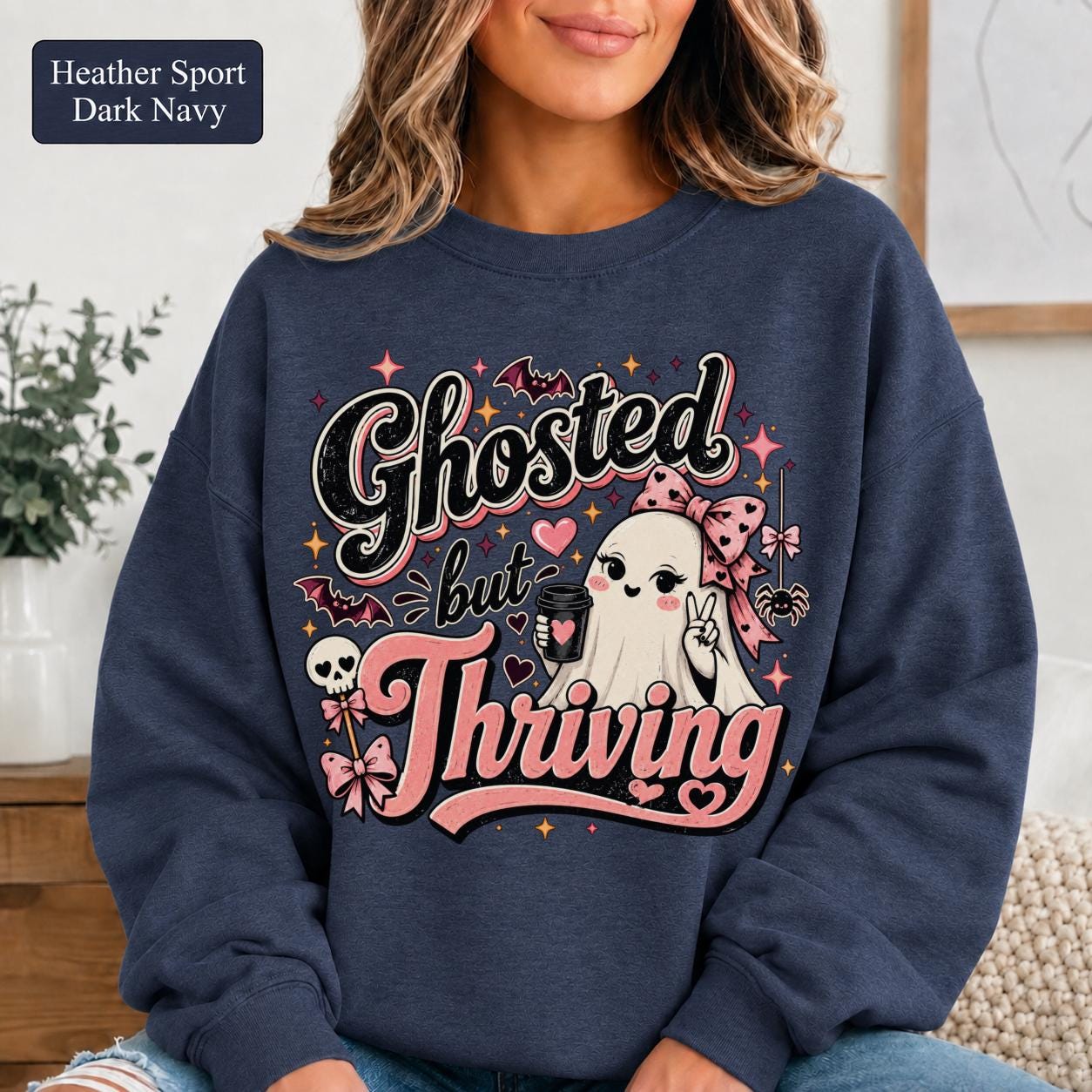 Woman wearing a heather sport dark navy crewneck sweatshirt featuring a cute bow-wearing ghost holding coffee, surrounded by pink hearts, bats, stars, and the words Ghosted but Thriving.