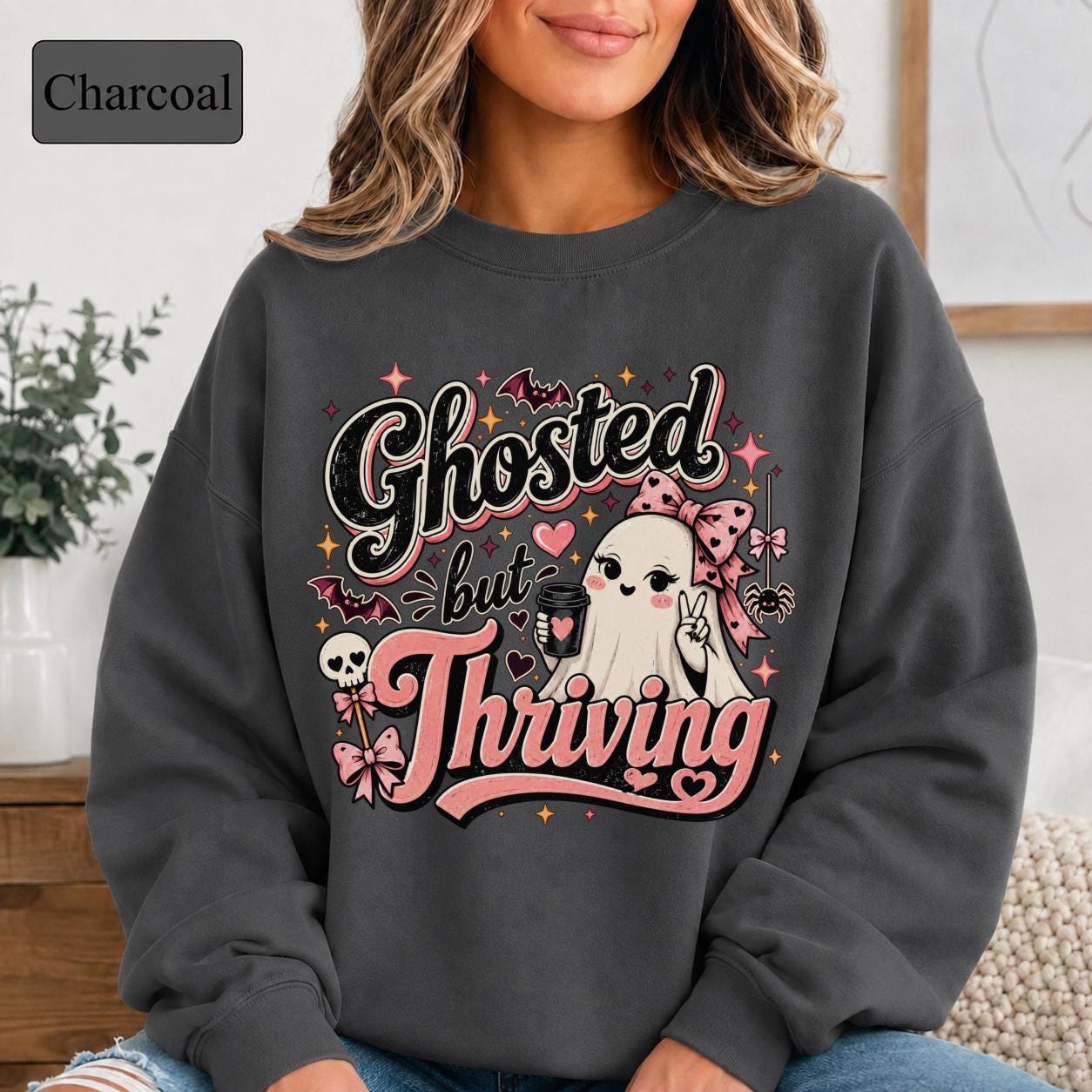 Woman wearing a charcoal crewneck sweatshirt featuring a cute bow-wearing ghost holding coffee, surrounded by pink hearts, bats, stars, and the words Ghosted but Thriving.