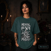 Mental Health Matters blue spruce T-shirt featuring a seated skeleton, flowers, butterflies, and bold gothic lettering in a monochrome design.