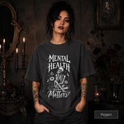 Mental Health Matters pepper T-shirt featuring a seated skeleton, flowers, butterflies, and bold gothic lettering in a monochrome design.