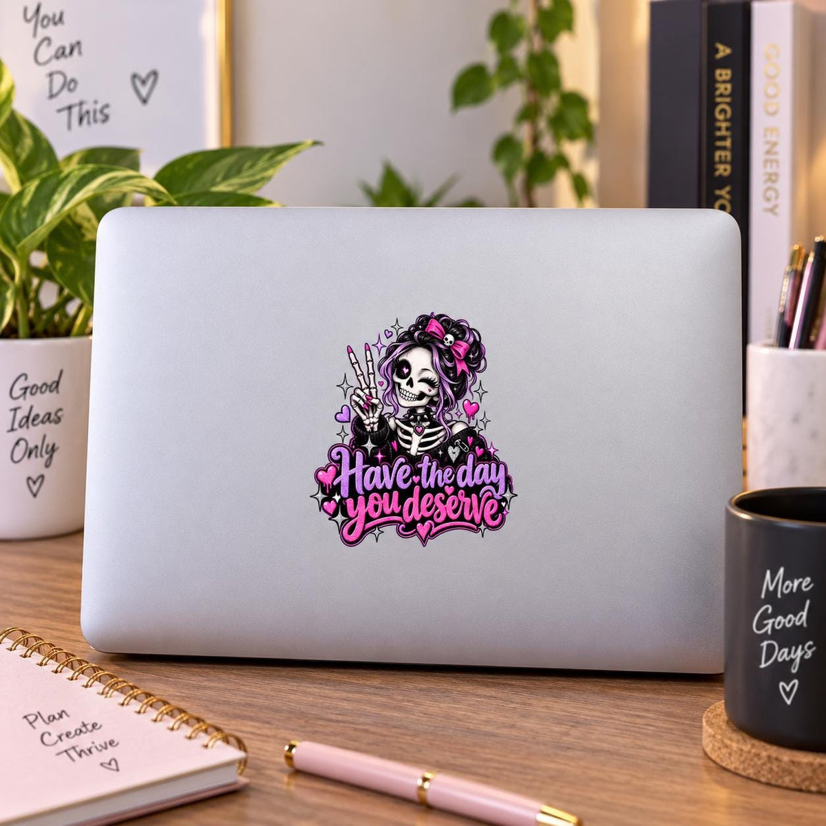 Have the Day You Deserve skeleton sticker shown on a silver laptop in a bright workspace with cute goth pink and purple details.