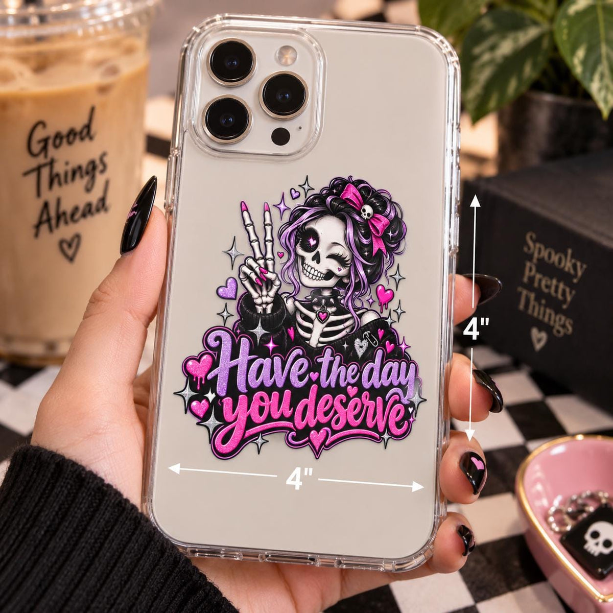 Have the Day You Deserve gothic skeleton decal displayed on a clear phone case with pink and purple pastel goth artwork.