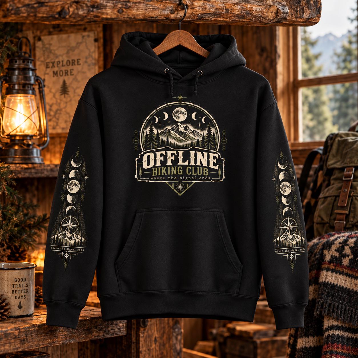 Black Offline Hiking Club hoodie hanging in a cozy rustic cabin, showing the chest design, kangaroo pocket, hood, and sleeve graphics.