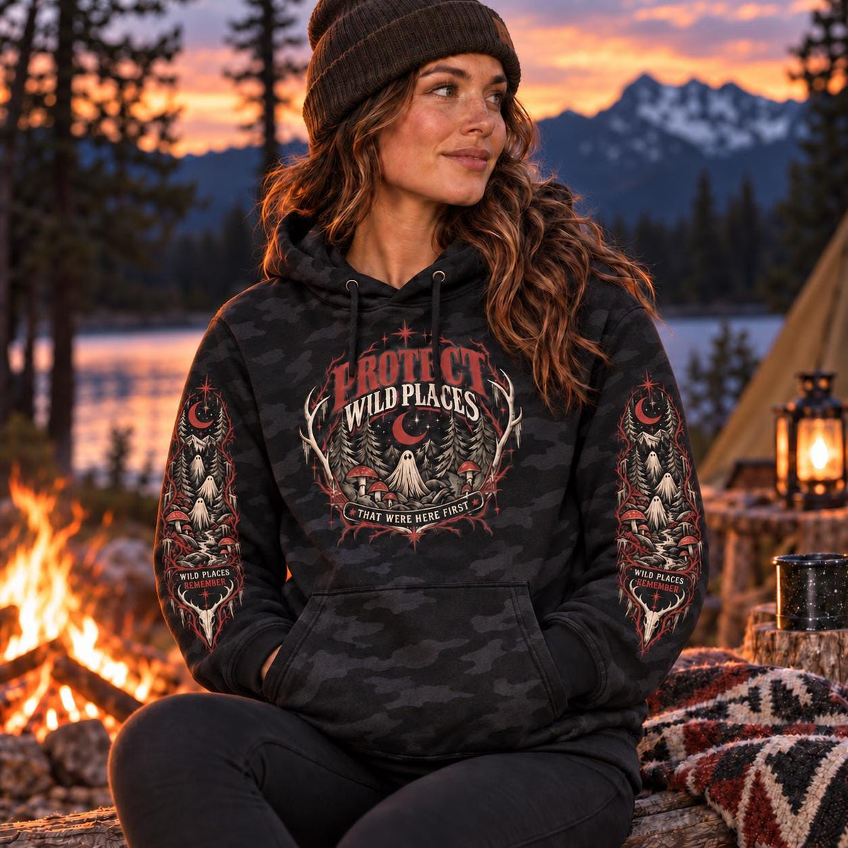 Woman wearing a black camo Protect Wild Places hoodie by a campfire at sunset, featuring gothic forest artwork on the chest and matching sleeve designs with ghosts, mushrooms, trees, and moon details.