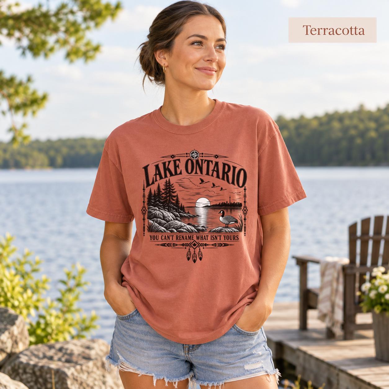 Terracotta Comfort Colors Lake Ontario T-shirt featuring a black vintage shoreline design with pine trees, rocky waterfront, Canada goose, Indigenous-inspired geometric details, and the phrase You Can’t Rename What Isn’t Yours.