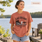 Terracotta Comfort Colors Lake Ontario T-shirt featuring a black vintage shoreline design with pine trees, rocky waterfront, Canada goose, Indigenous-inspired geometric details, and the phrase You Can’t Rename What Isn’t Yours.