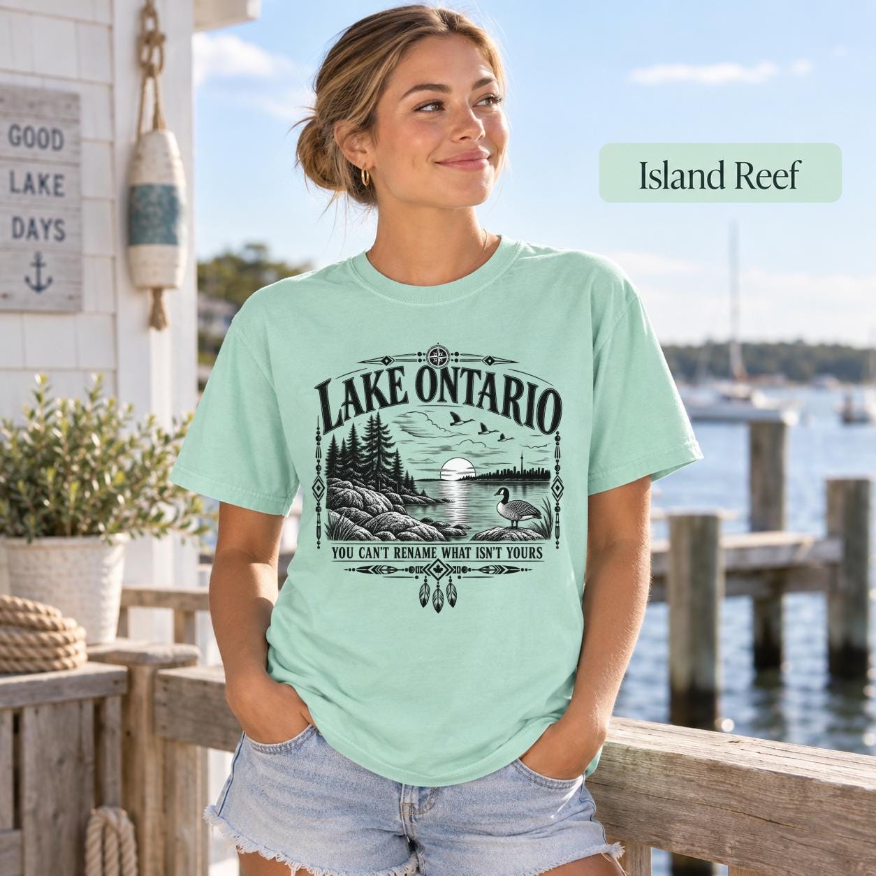Island Reef Comfort Colors Lake Ontario T-shirt featuring a black vintage shoreline design with pine trees, rocky waterfront, Canada goose, Indigenous-inspired geometric details, and the phrase You Can’t Rename What Isn’t Yours.