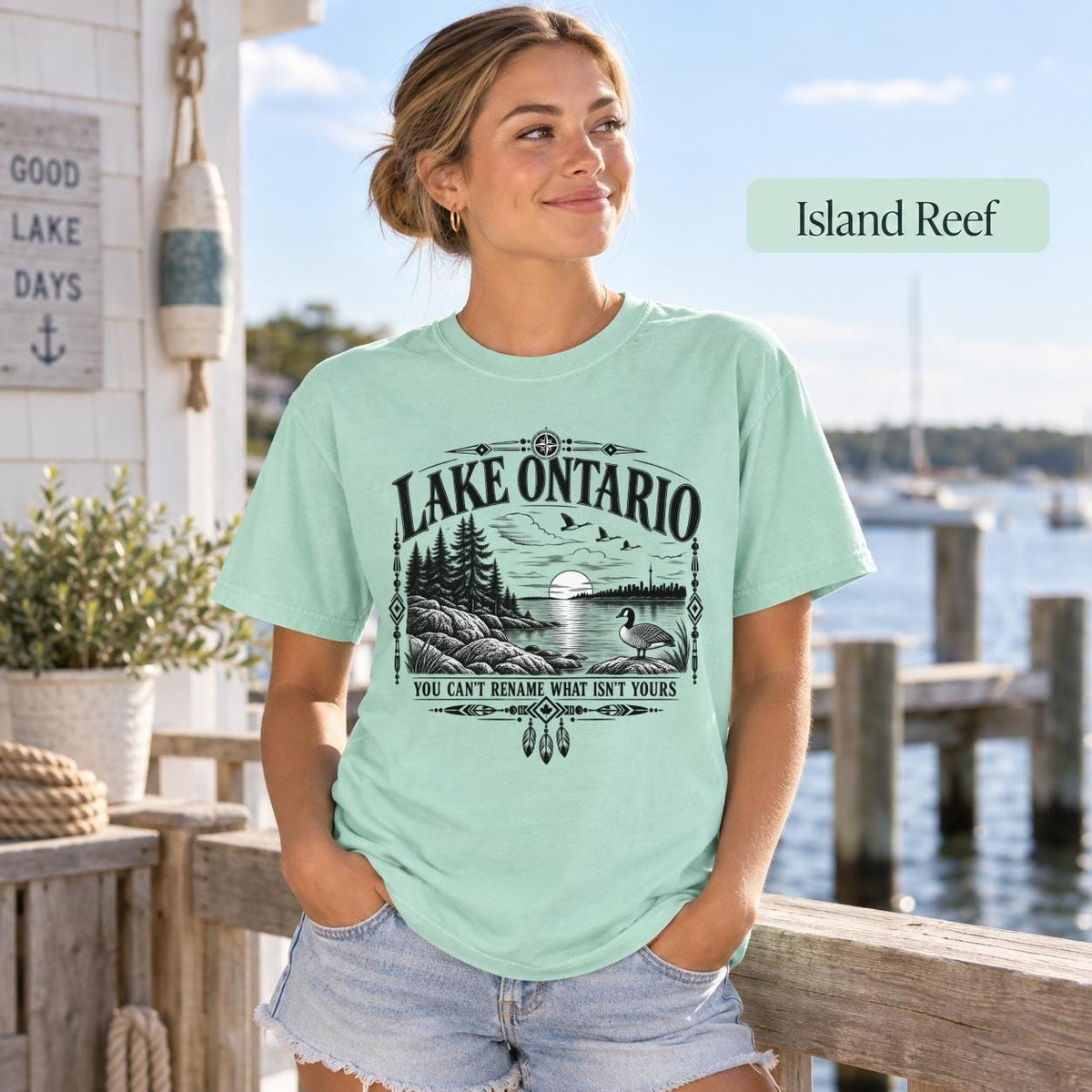 Island Reef Comfort Colors Lake Ontario T-shirt featuring a black vintage shoreline design with pine trees, rocky waterfront, Canada goose, Indigenous-inspired geometric details, and the phrase You Can’t Rename What Isn’t Yours.