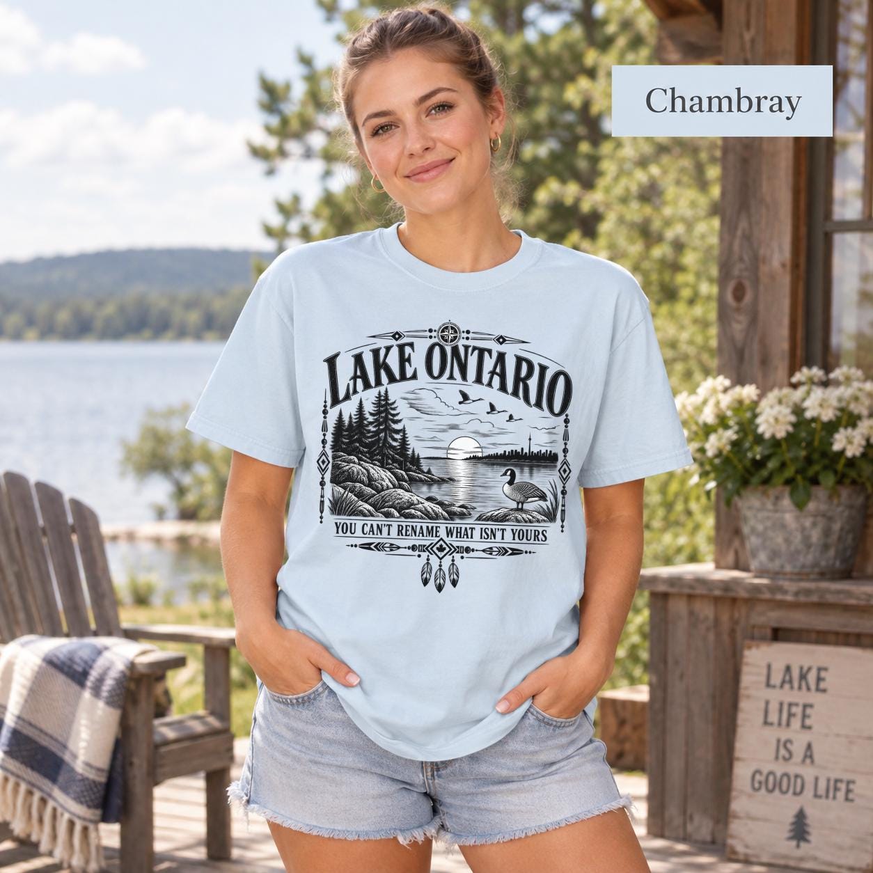Chambray Comfort Colors Lake Ontario T-shirt featuring a black vintage shoreline design with pine trees, rocky waterfront, Canada goose, Indigenous-inspired geometric details, and the phrase You Can’t Rename What Isn’t Yours.