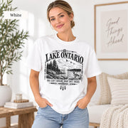 White Comfort Colors Lake Ontario T-shirt featuring a black vintage shoreline design with pine trees, rocky waterfront, Canada goose, Indigenous-inspired geometric details, and the phrase You Can’t Rename What Isn’t Yours.
