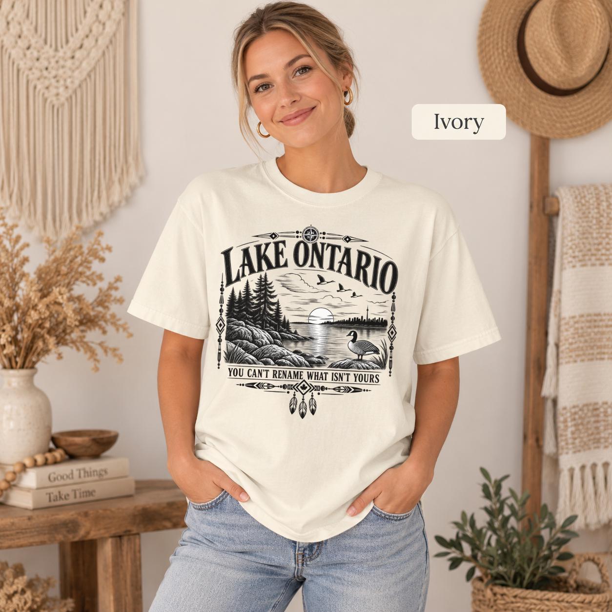 Ivory Comfort Colors Lake Ontario T-shirt featuring a black vintage shoreline design with pine trees, rocky waterfront, Canada goose, Indigenous-inspired geometric details, and the phrase You Can’t Rename What Isn’t Yours.
