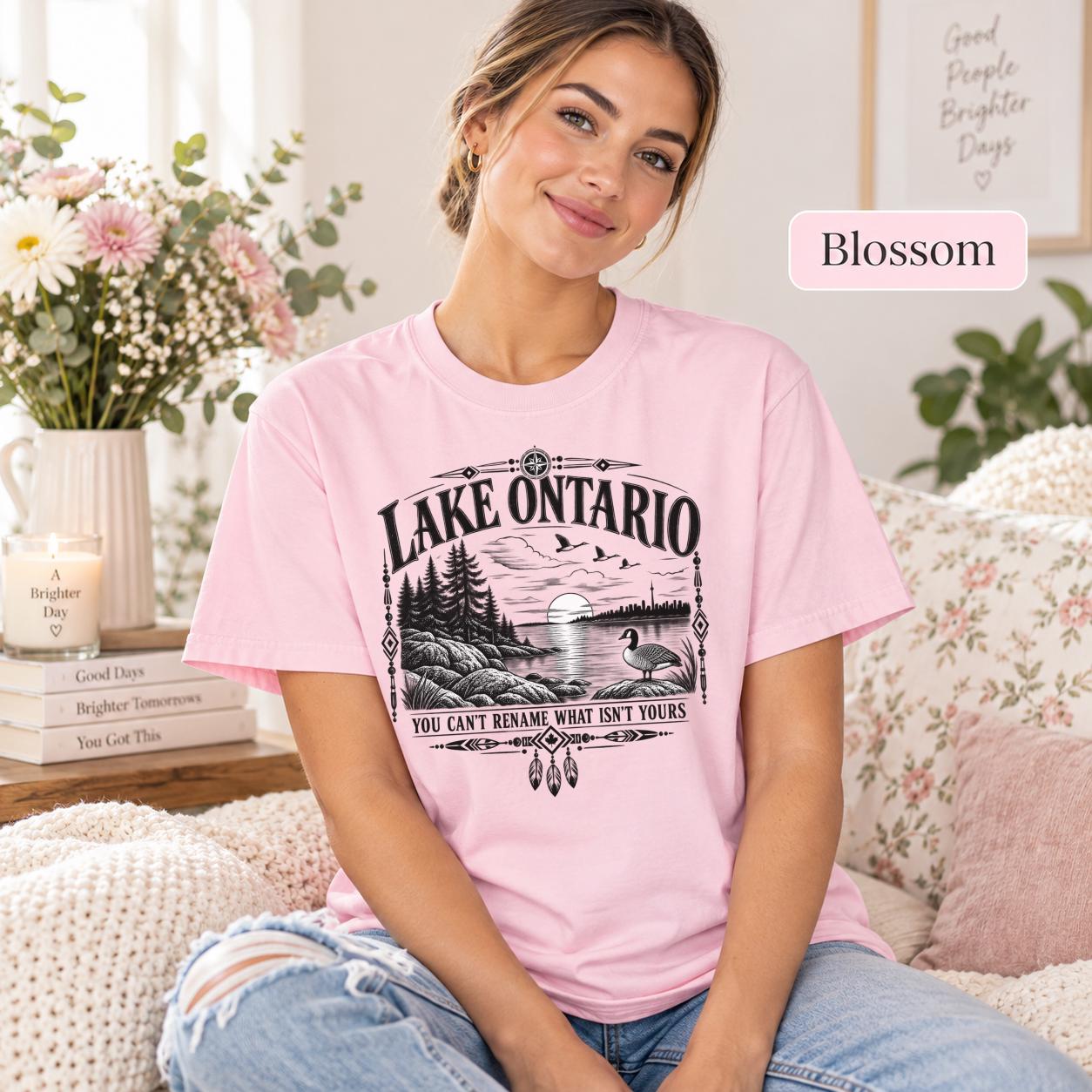 Blossom Comfort Colors Lake Ontario T-shirt featuring a black vintage shoreline design with pine trees, rocky waterfront, Canada goose, Indigenous-inspired geometric details, and the phrase You Can’t Rename What Isn’t Yours.