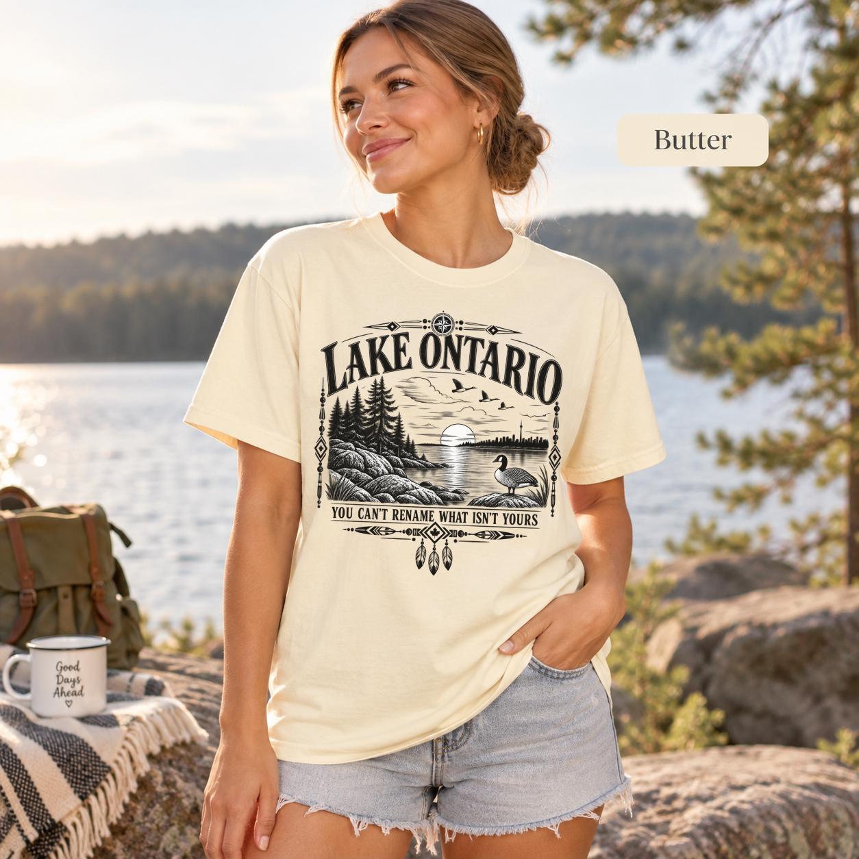 Butter Comfort Colors Lake Ontario T-shirt featuring a black vintage shoreline design with pine trees, rocky waterfront, Canada goose, Indigenous-inspired geometric details, and the phrase You Can’t Rename What Isn’t Yours.