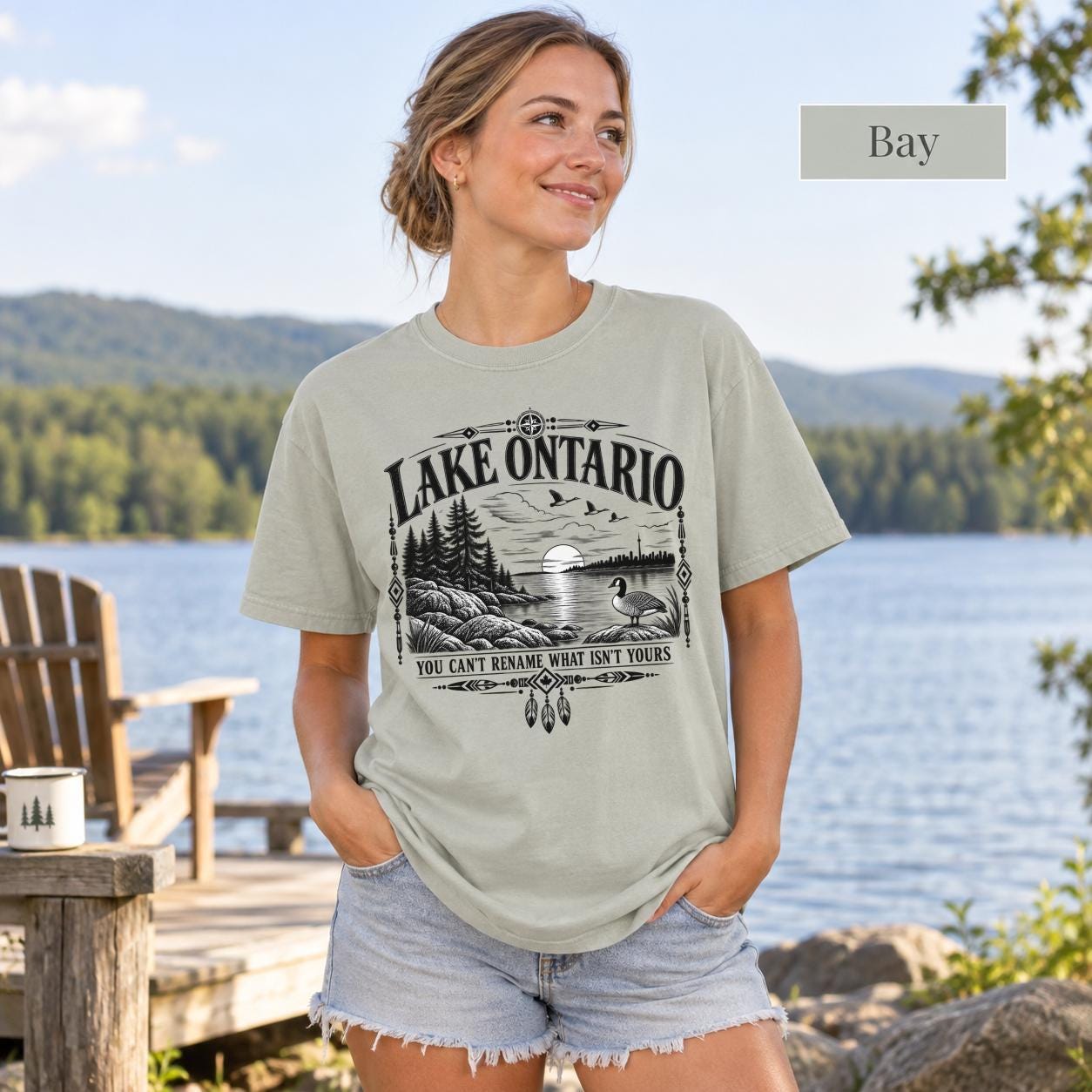 Bay Comfort Colors Lake Ontario T-shirt featuring a black vintage shoreline design with pine trees, rocky waterfront, Canada goose, Indigenous-inspired geometric details, and the phrase You Can’t Rename What Isn’t Yours.