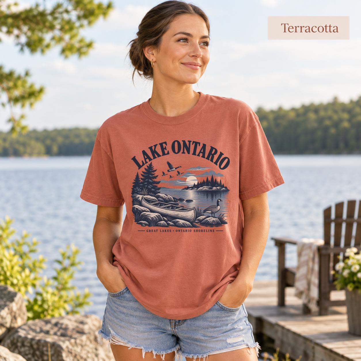 Woman wearing a Terracotta Comfort Colors T-shirt featuring a vintage Lake Ontario shoreline graphic with canoe, pine trees, rocky shore, Canada geese, water, and Great Lakes Ontario Shoreline text.