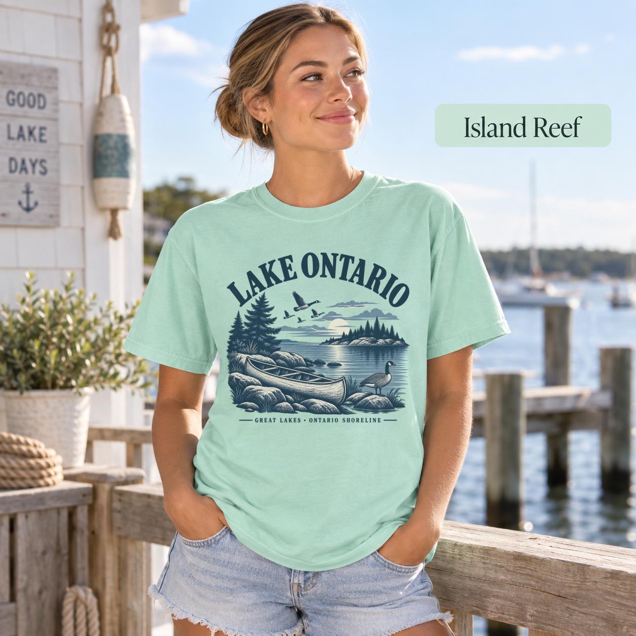 Woman wearing a Island Reef Comfort Colors T-shirt featuring a vintage Lake Ontario shoreline graphic with canoe, pine trees, rocky shore, Canada geese, water, and Great Lakes Ontario Shoreline text.