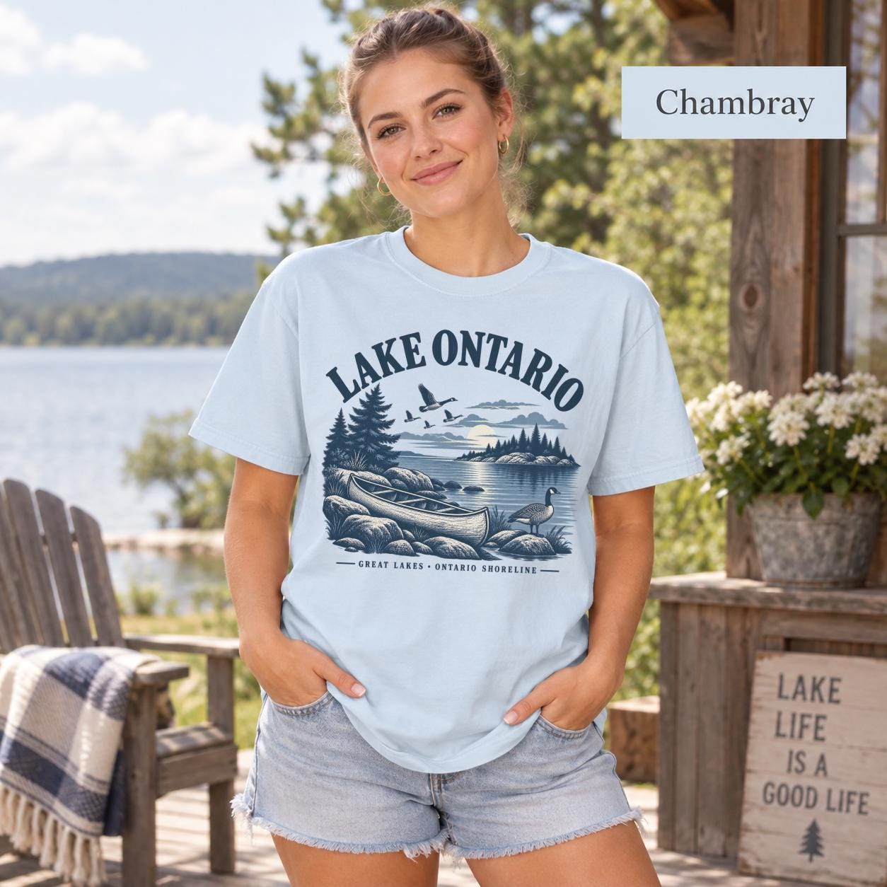 Woman wearing a Chambray Comfort Colors T-shirt featuring a vintage Lake Ontario shoreline graphic with canoe, pine trees, rocky shore, Canada geese, water, and Great Lakes Ontario Shoreline text.