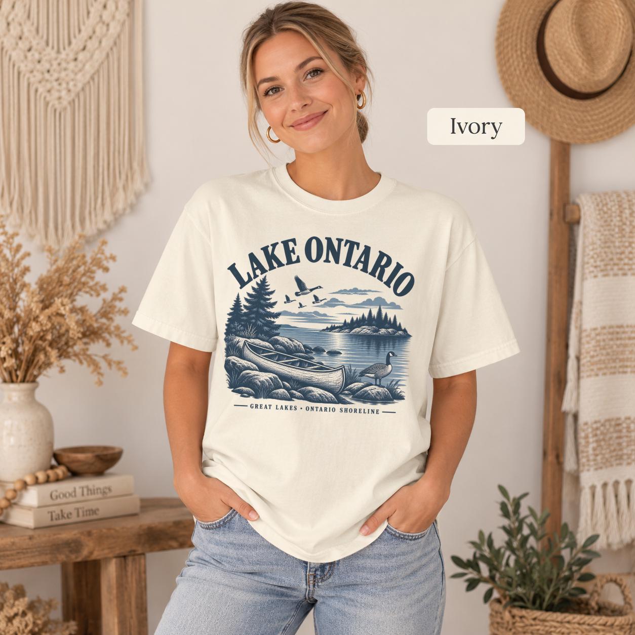 Woman wearing a Ivory Comfort Colors T-shirt featuring a vintage Lake Ontario shoreline graphic with canoe, pine trees, rocky shore, Canada geese, water, and Great Lakes Ontario Shoreline text.