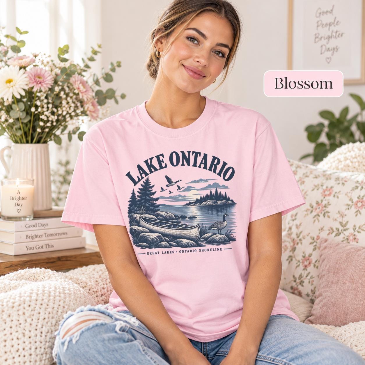 Woman wearing a Blossom Comfort Colors T-shirt featuring a vintage Lake Ontario shoreline graphic with canoe, pine trees, rocky shore, Canada geese, water, and Great Lakes Ontario Shoreline text.